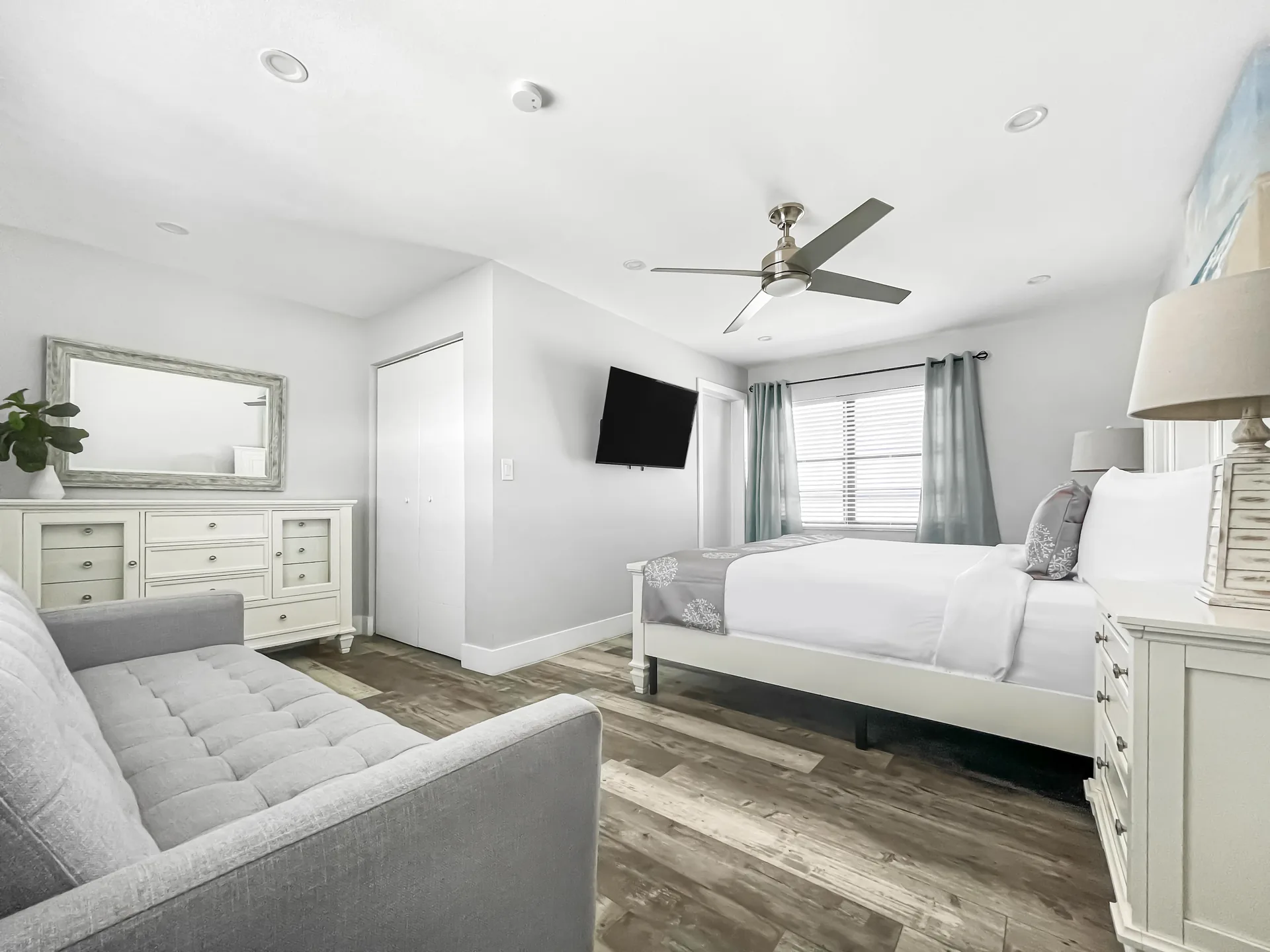 A wide view of a hotel room with a white bed, dresser, TV, sofa, and ceiling fan.