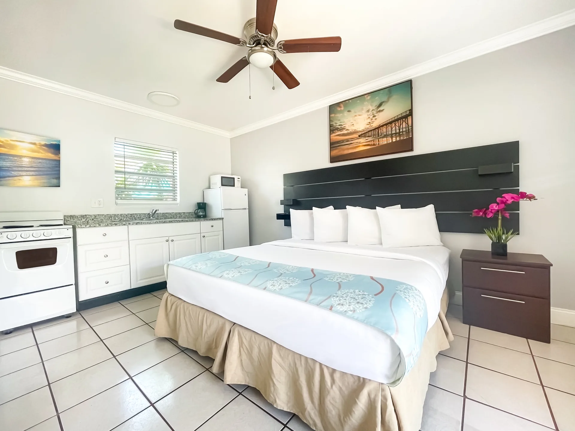 A hotel room with a double bed, kitchenette, and ceiling fan.