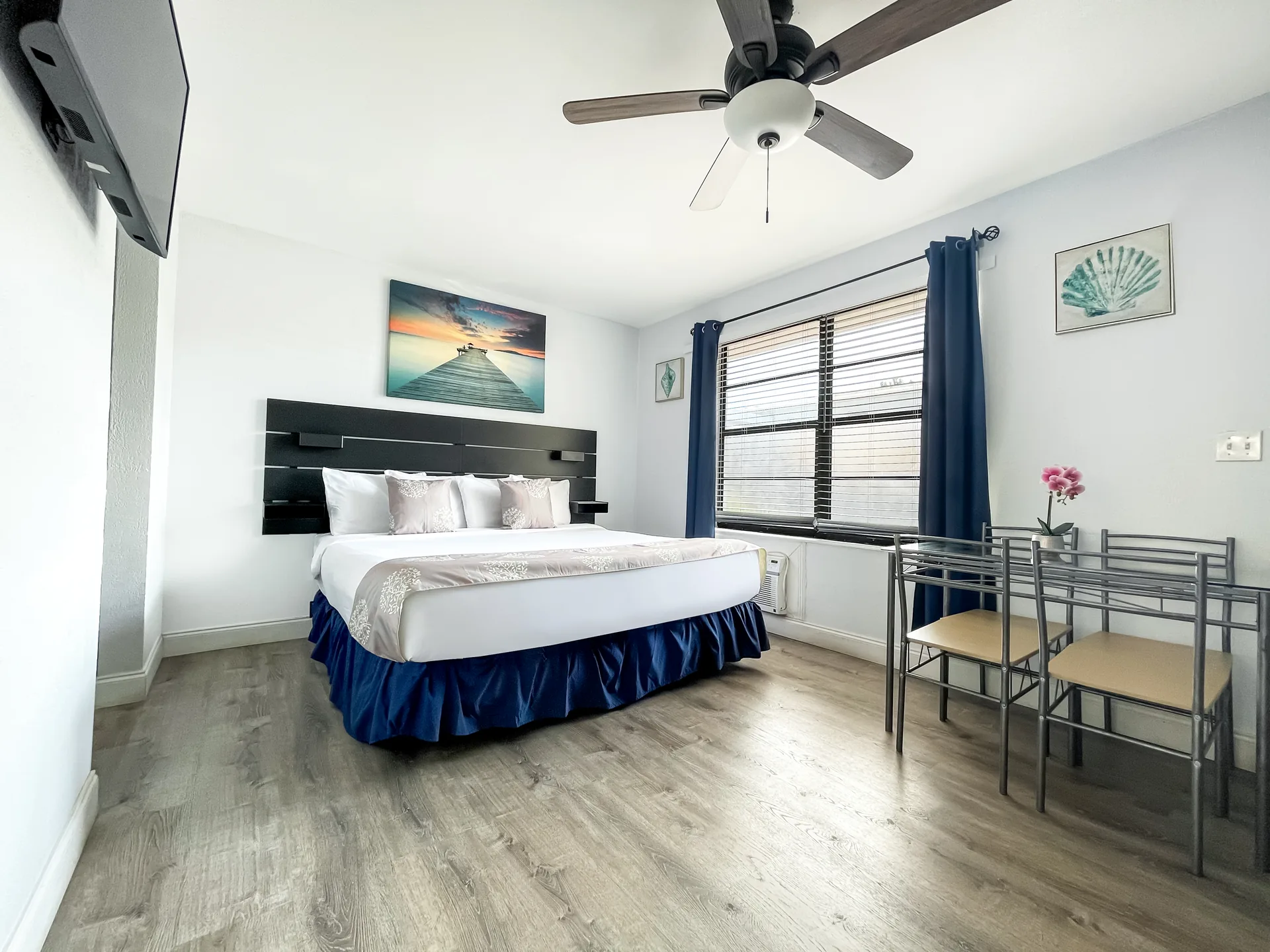A hotel room with a bed, dining table, ceiling fan, and window